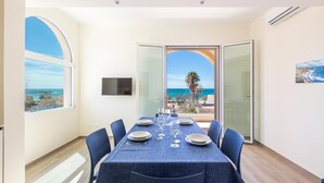 Ein Esszimmer mit einem für sechs Personen gedeckten Tisch, großen Fenstern mit Blick auf das Meer und einem Gemälde an der Wand.