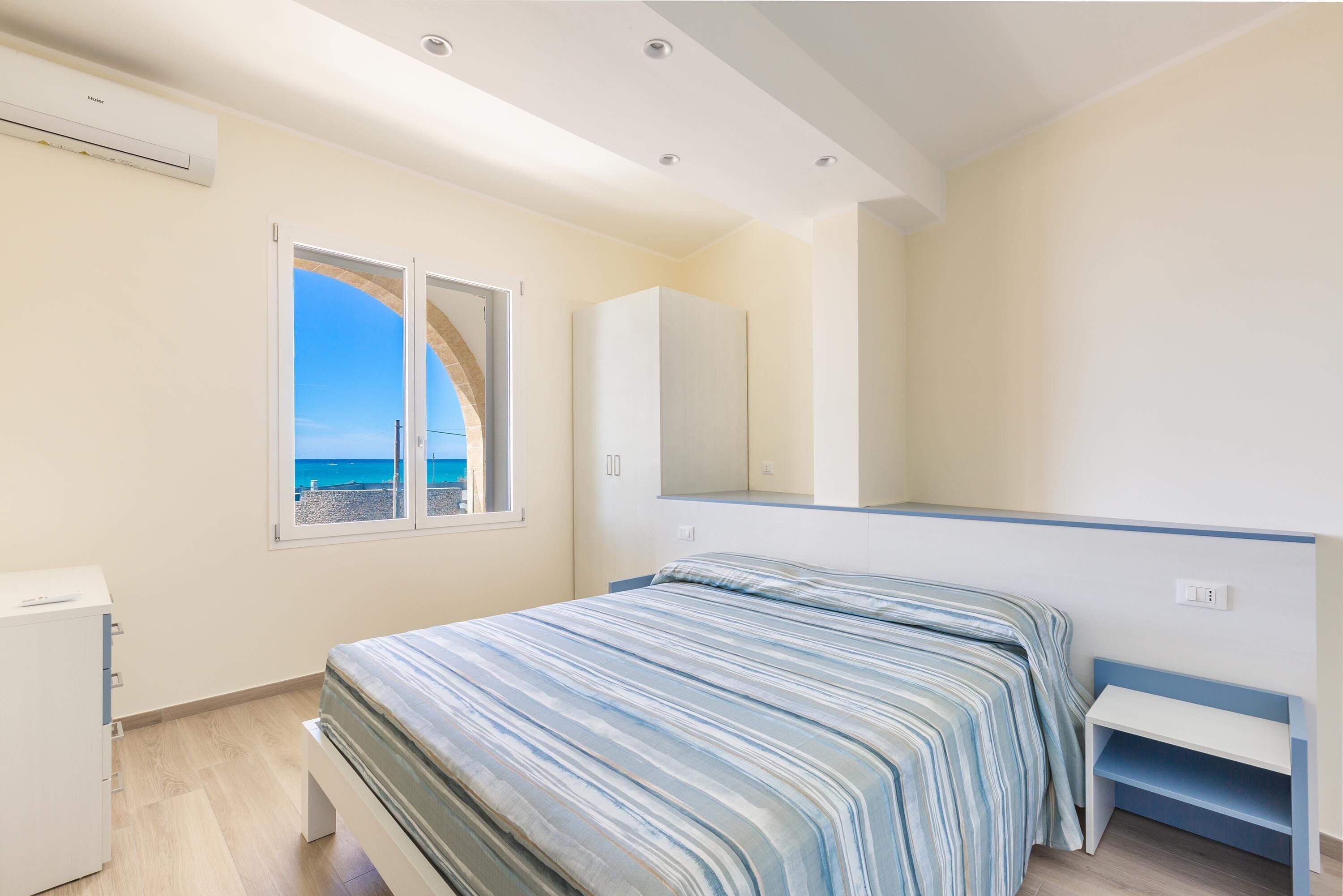 A bedroom with a bed, a wardrobe, a desk, and a window with a view of the sea.
