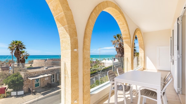Ein Balkon mit weißem Tisch und Stühlen, der Blick auf die Küste mit Palmen und einem strahlend blauen Himmel gewährt.