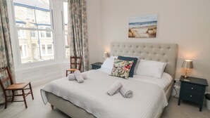 A neatly made bed with pillows and a rolled towel, a chair, a bedside table with a lamp, and a painting on the wall.