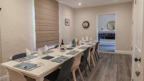 A dining area with a long table set for six, chairs, a clock, and a kitchen visible through an open door.