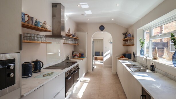A modern kitchen with a central island, built-in oven, and stainless steel appliances. There are wooden shelves with kitchenware and a window with a view.