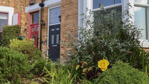 A brick house with a black door, a red door, and a window with blinds. There is a yellow rose in the garden.