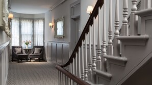 A hallway with a carpeted floor, two armchairs with cushions, a small table with a vase of flowers, and a staircase with white balusters. - Carroll Villa Hotel (Cape May)