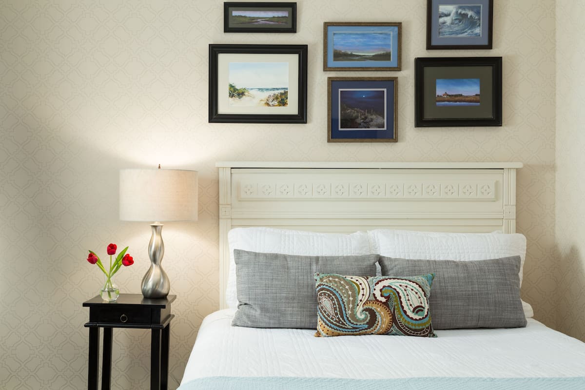 A neatly arranged bedroom with a bed, pillows, a nightstand with a vase of flowers, and framed pictures on the wall.