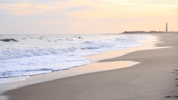 A beach with waves, a sandy shore, and a lighthouse in the distance. - Carroll Villa Hotel (Cape May)