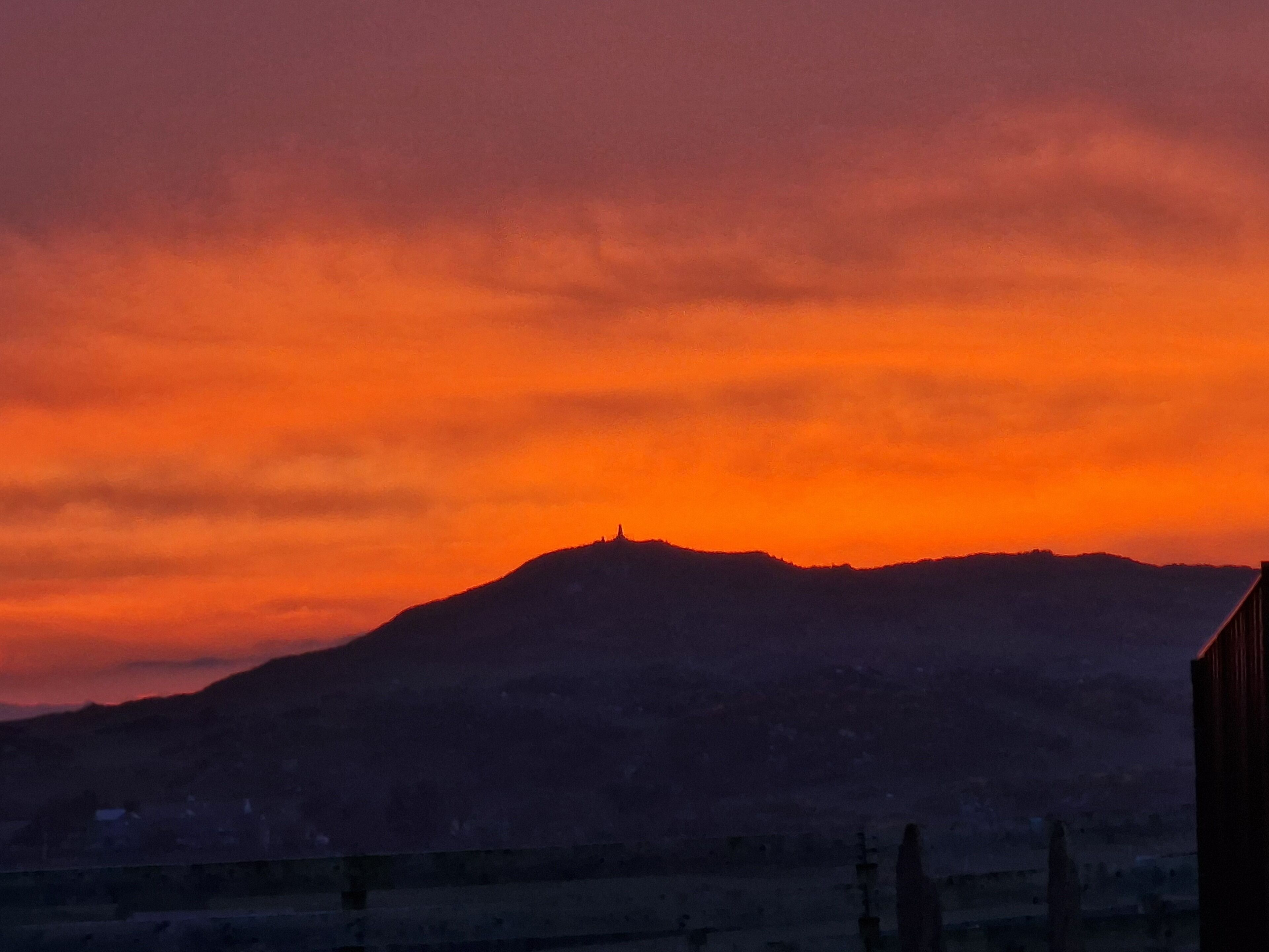 A sunset over a mountainous landscape with a silhouette of a hill and a fence in the foreground.