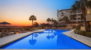 Relax by the resort's rectangular pool surrounded by lounge chairs and umbrellas, with palm trees and modern buildings in the background.