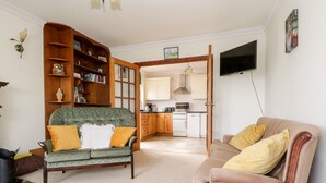 A living room with a green sofa, a brown sofa, a wooden bookshelf, and a kitchen in the background.