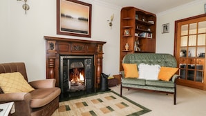 Een woonkamer met een open haard, een groene bank met oranje kussens, een bruine leren fauteuil en een houten boekenkast.