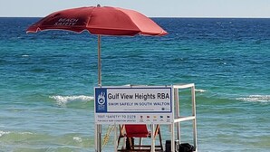 Ein Rettungsschwimmerstand mit einem roten Sonnenschirm und einem Schild für Gulf View Heights RBA, das darauf hinweist, eine SMS mit dem Wort „SICHERHEIT“ für aktuelle Informationen zu senden.