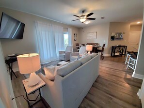 Een woonkamer met een bank, twee fauteuils, een salontafel en een aan de muur bevestigde televisie. Er is een plafondventilator, een eethoek met een tafel en stoelen, en een bijzettafeltje met een lamp.