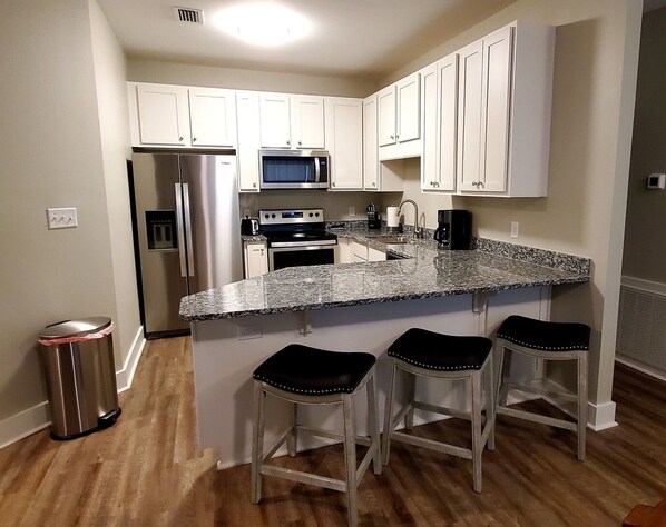 A kitchen with white cabinets, stainless steel appliances, a granite countertop, and three bar stools.