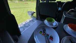 A van interior with a table set for a meal, a portable grill, and a scenic view of a green landscape. - Inviting 2 bed Camper in Holyhead (Holyhead)
