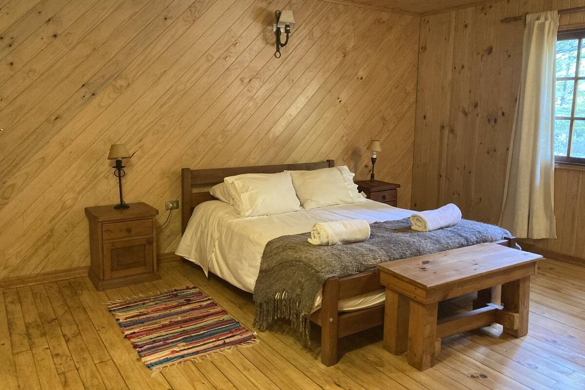 A cozy bedroom with a wooden sloped ceiling, a bed with white linens, two bedside tables with lamps, a wooden bench, and a colorful rug.