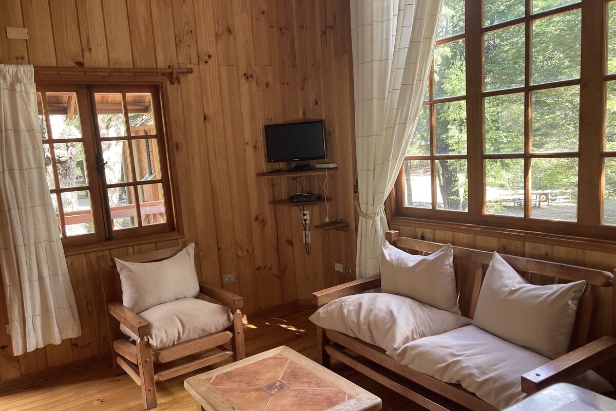 A wooden cabin interior with a sofa, two chairs, a small table, a television, and large windows with curtains.