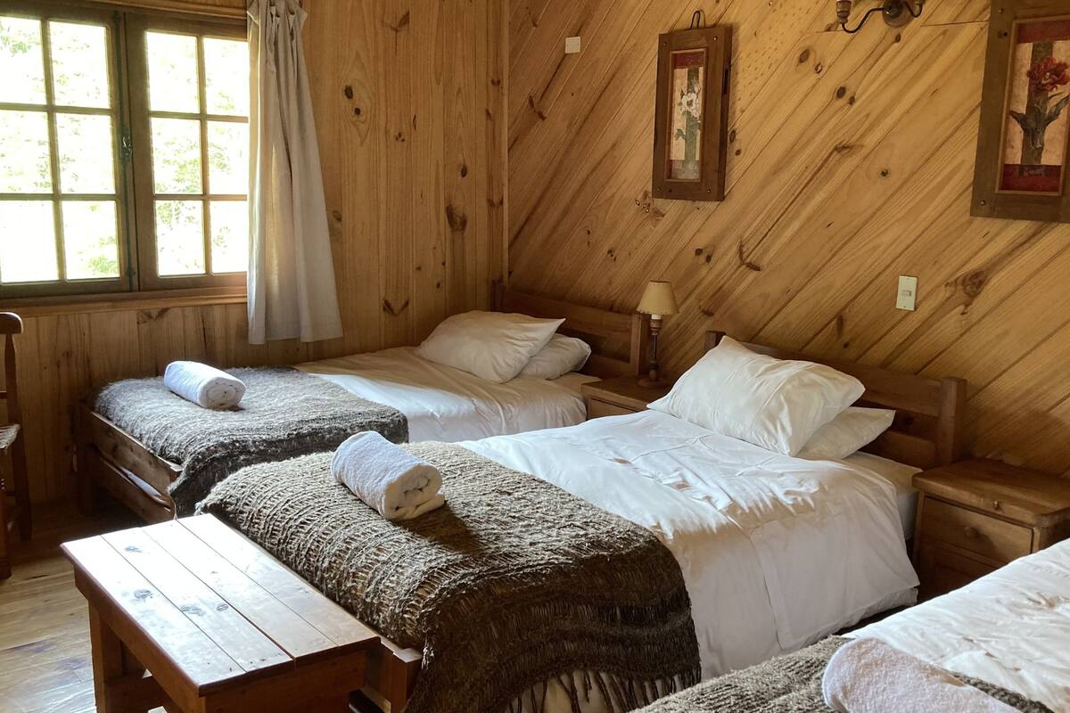 A rustic wooden room with three beds, each with white linens and rolled towels. There is a wooden bench and a table, and the room has wooden walls with two framed pictures.