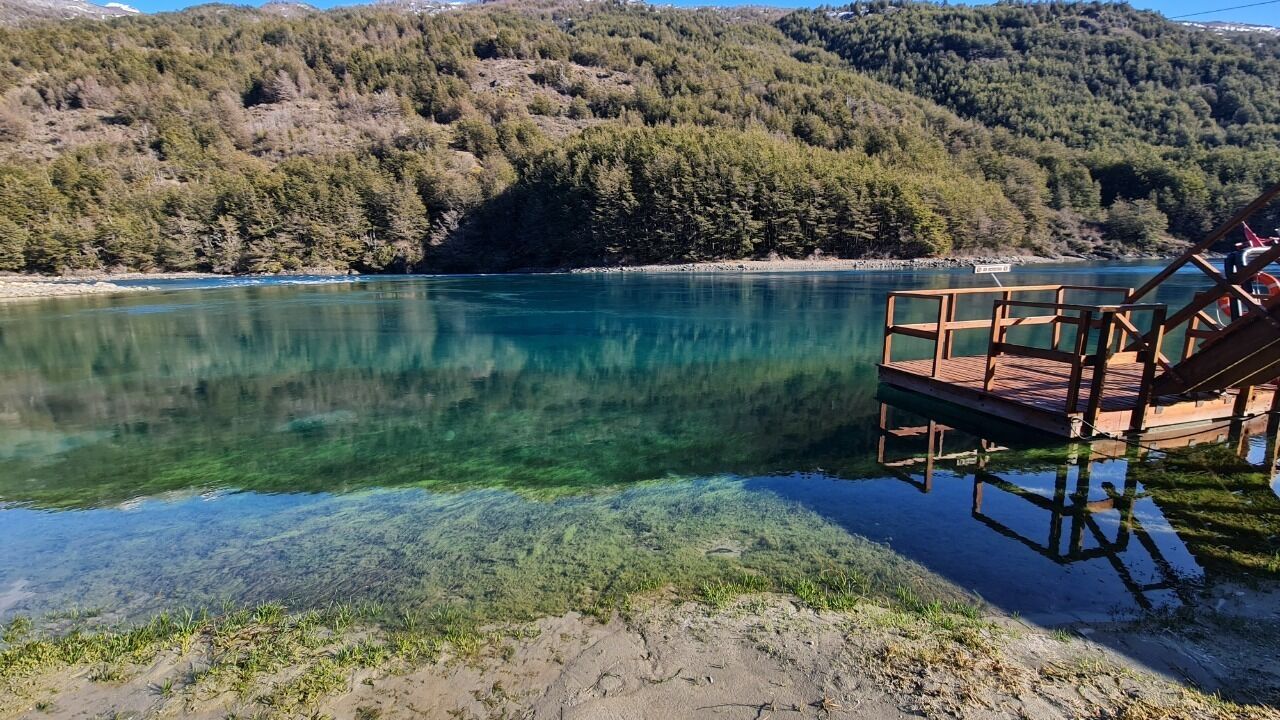 A clear lake with a wooden dock and a chair, surrounded by forested hills.
