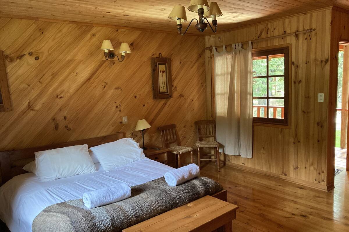 A wooden cabin bedroom with a bed, two chairs, a bench, a window with curtains, and a chandelier.
