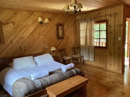A wooden cabin bedroom with a bed, two chairs, a bench, a window with curtains, and a chandelier.