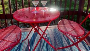Mesa metálica roja con dos sillas y dos copas de martini en una terraza con barandilla de madera, rodeada de árboles.