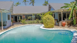 A pool area with a large decorative plant, a covered seating area, and a building with a tiled roof. - Seven Palms Villa  (Runaway Bay)