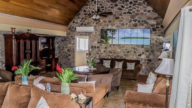 A cozy living room with stone walls, a wooden ceiling, and a large window overlooking a scenic view. - Seven Palms Villa  (Runaway Bay)