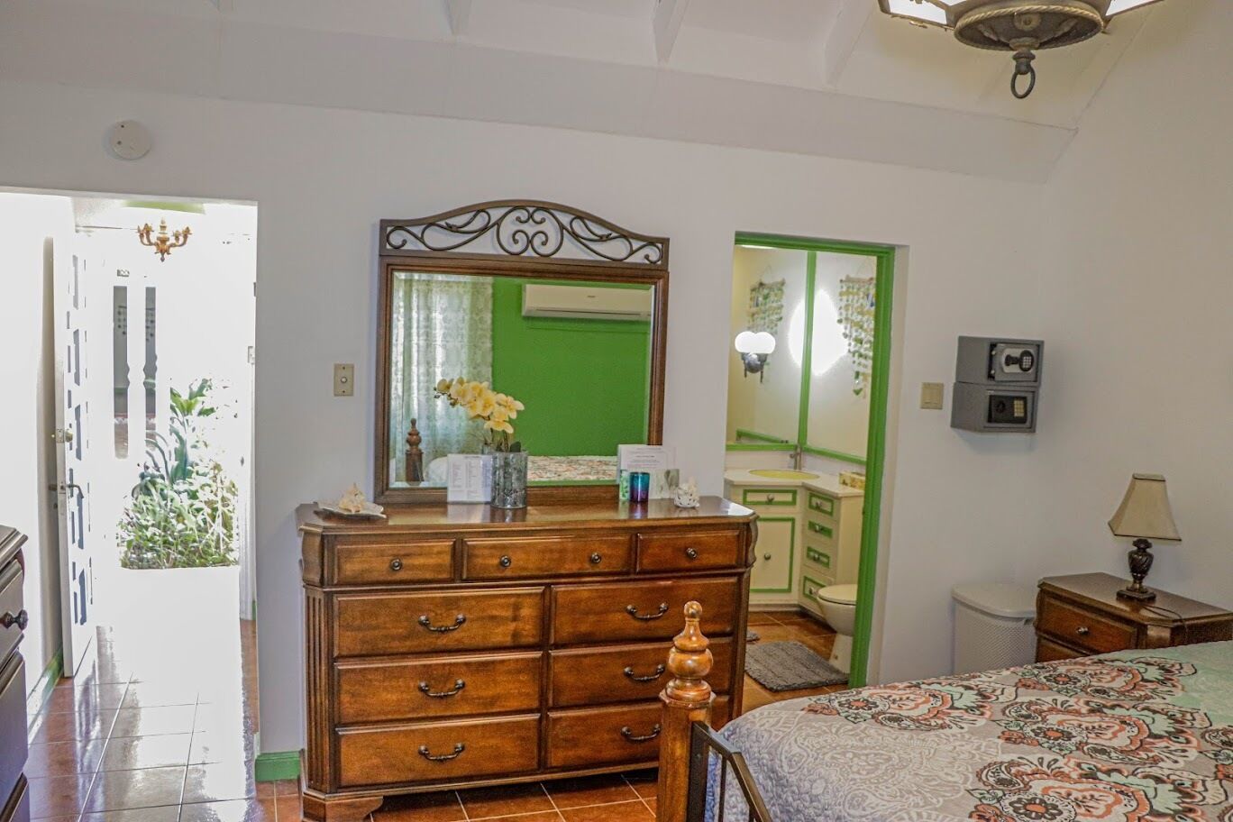A bedroom with a wooden dresser, a bed with a patterned bedspread, a green door, and a small bedside table with a lamp.
