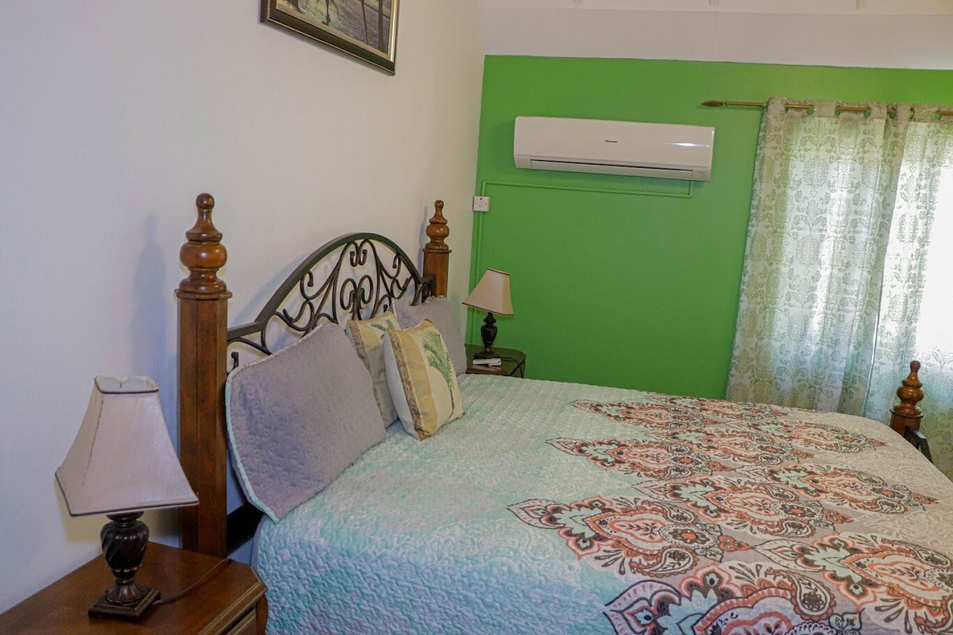 Un dormitorio con pared verde, colcha estampada, cabecero de madera, dos lámparas de noche y un aire acondicionado sobre la cama.
