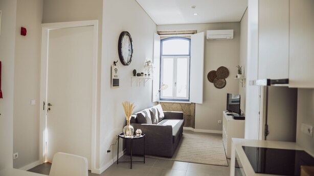 A modern living room with a grey sofa, a small side table with decorative items, a white chair, a large window with a view, and a flat-screen TV on a stand.