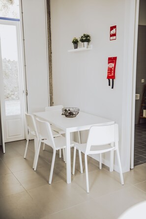Uma mesa de jantar branca com quatro cadeiras, uma pequena planta numa prateleira e um sinal de saída de incêndio na parede.