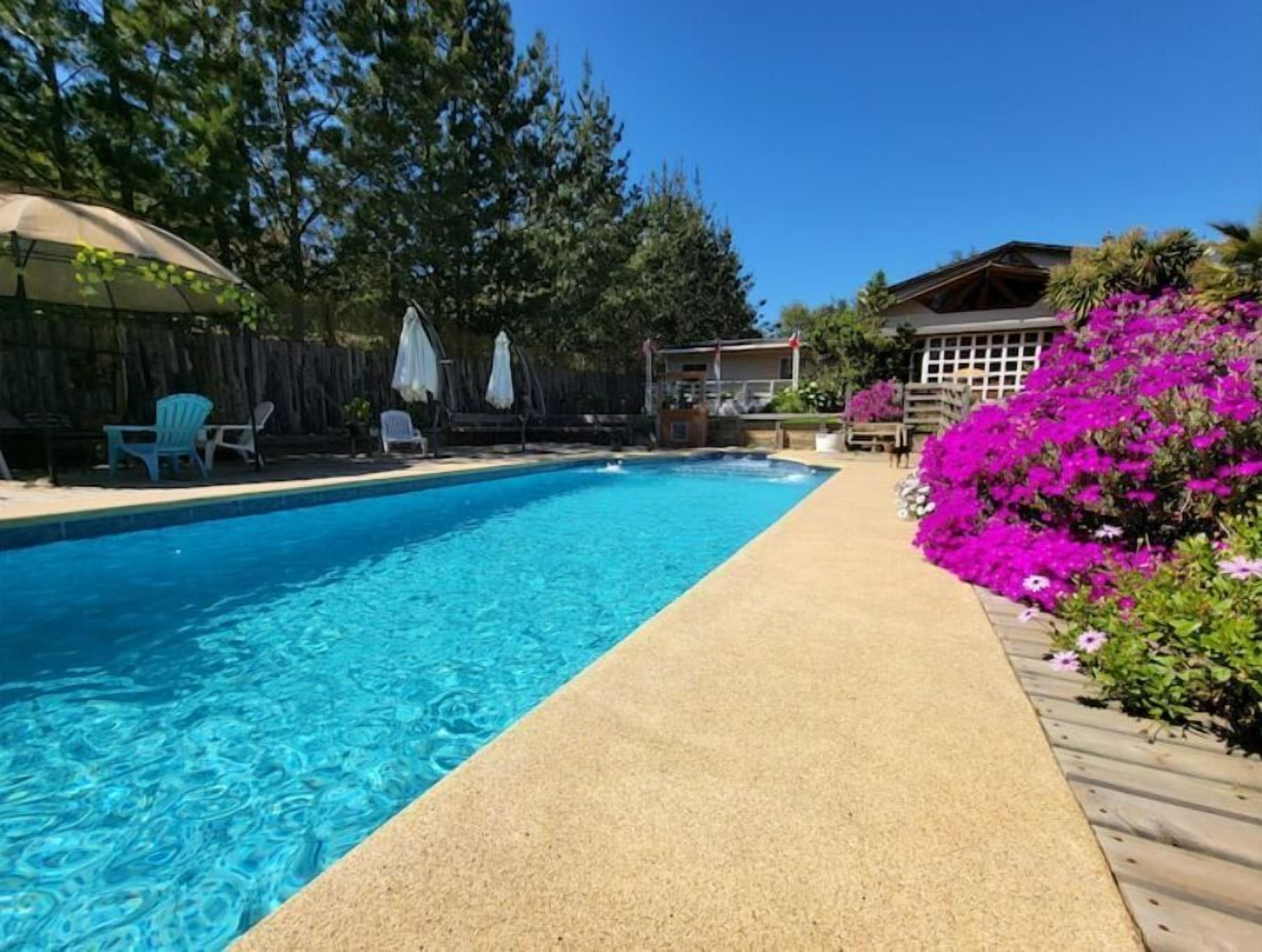 Ein Schwimmbecken mit klarem, blauem Wasser, umgeben von einer Holzterrasse und einem Garten mit rosa Blumen.