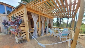 Una estructura de madera con un patio cubierto, que incluye una mesa y sillas, y una cerca de malla metálica.