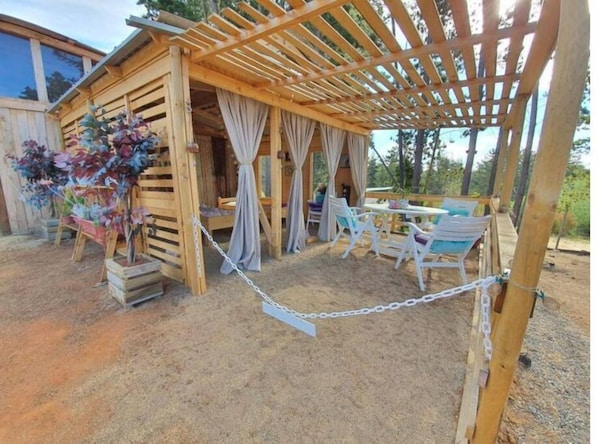 Una estructura de madera con un patio cubierto, que incluye una mesa y sillas, y una cerca de malla metálica.