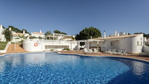 A swimming pool with clear blue water, white poolside chairs, and a white building with a tiled roof.