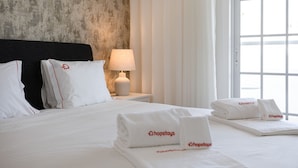A hotel room with a bed, white linens, a nightstand with a lamp, and a window with curtains.