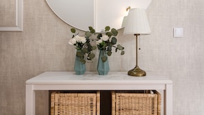 A white console table with two teal vases containing white flowers and green leaves, a brass floor lamp, and wicker baskets underneath.
