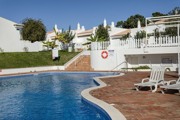 A swimming pool with a brick deck and lounge chairs, surrounded by white buildings with a white picket fence. - Vilamoura Eden Villas by Hopstays | Pool & Golf (Vilamoura)