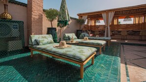 Un área junto a la piscina con dos sillones de descanso, una pérgola de madera y suelo de baldosas.