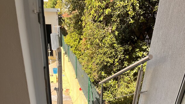 Vista de varanda com guarda-corrimão de metal, piso de azulejos e cerca verde ao fundo.