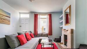 Una sala de estar moderna con un sofá gris, cojines rojos, una mesa de centro de vidrio, una alfombra roja, una chimenea y un ventanal con cortinas rojas.