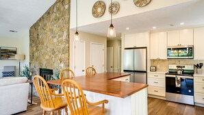 A modern kitchen with white cabinets, a large island with wooden chairs, a stone accent wall, and a dining area with a sofa and lamp. - Charming Condo on Sky Valley Golf Course! (Sky Valley)