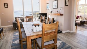 Un'area pranzo con tavolo e sedie in legno, una televisione e vista sul mare attraverso ampie finestre.