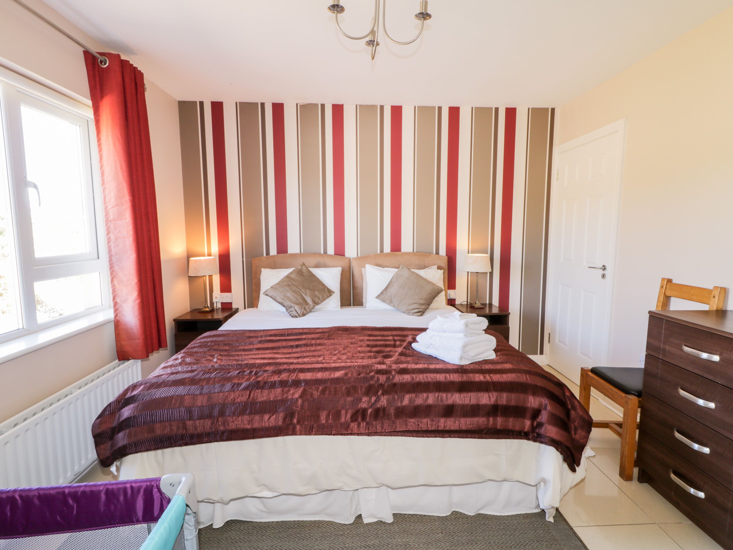 A hotel room with a large bed, striped wallpaper, a chandelier, a wooden dresser, and a window with curtains.