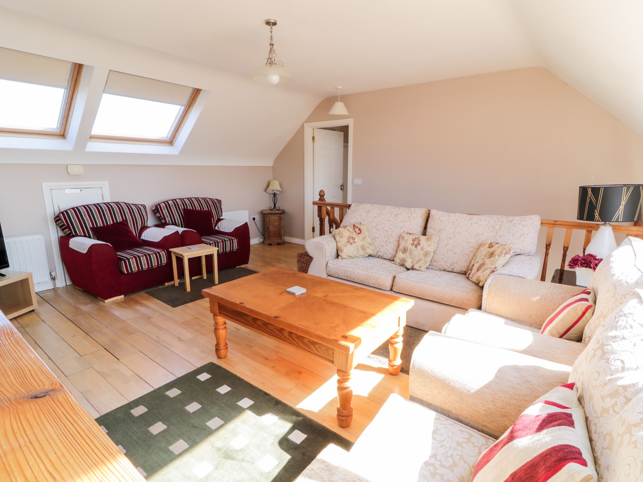 A cozy living room with a sofa, armchairs, a coffee table, and a TV. There are two windows providing natural light.