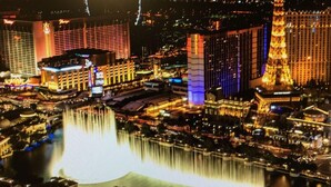 A nighttime view of a cityscape with illuminated buildings, a large fountain, and a prominent tower.