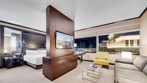 A modern hotel room with a large bed, a desk, a yellow armchair, a grey sofa, and a city view through large windows.