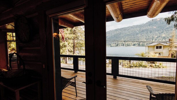A wooden cabin with a deck overlooking a lake and mountains.