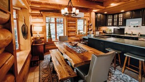 A log cabin interior with a wooden dining table, benches, a chandelier, and a kitchen area.