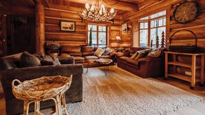 A cozy log cabin interior with a fireplace, leather sofas, a chandelier, and a large window offering a view of the forest.